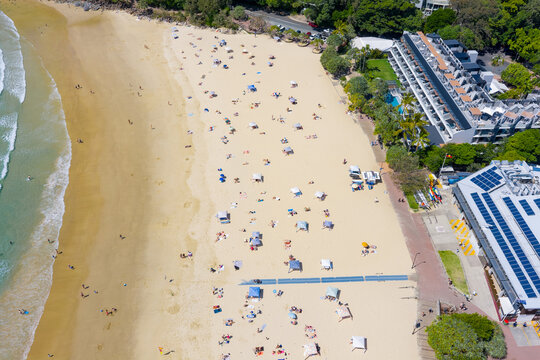 Aerial view of bathers spread over a wide sandy beach below rows of waterfront accommodation
