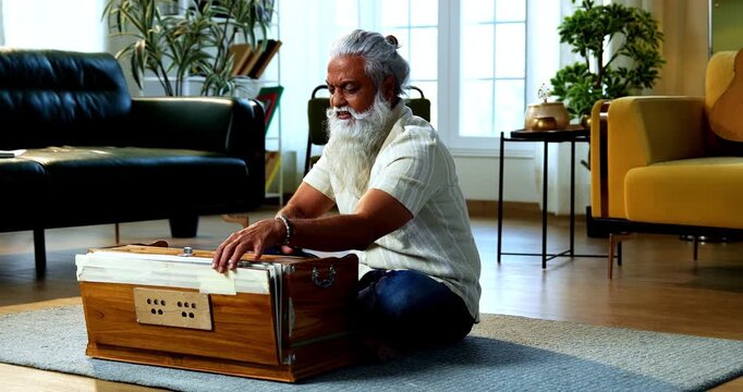 Indian mid age stylish bearded man practicing vocals or singing at home, dedicated to daily riyaz while accompanying harmonium in modern home studio interior setting