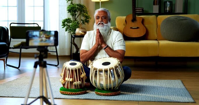 Indian mid age stylish bearded man taking online Tabla musical instrument playing class at home, experienced classical percussion tutor streaming lesson on smartphone in modern studio