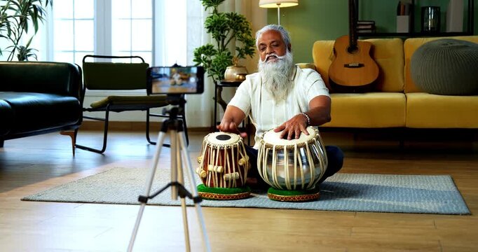 Indian mid age stylish bearded man taking online Tabla musical instrument playing class at home, experienced classical percussion tutor streaming lesson on smartphone in modern studio