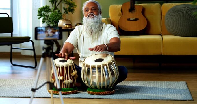 Indian mid age stylish bearded man taking online Tabla musical instrument playing class at home, experienced classical percussion tutor streaming lesson on smartphone in modern studio