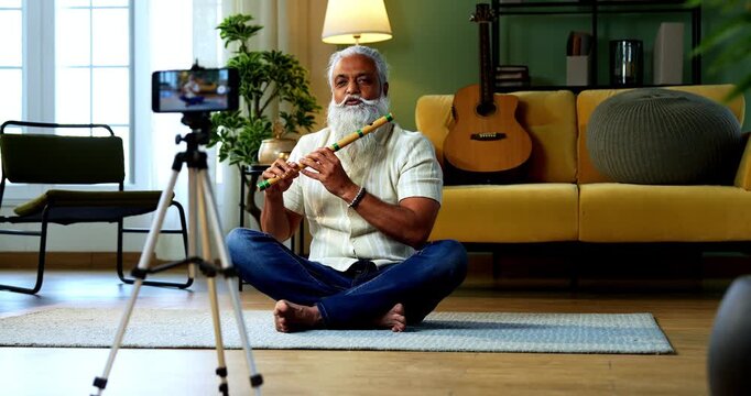 Indian mid age stylish bearded man teaching playing wooden flute musical instrument at home using smartphone, experienced classical bansuri tutor conducting online class in modern studio
