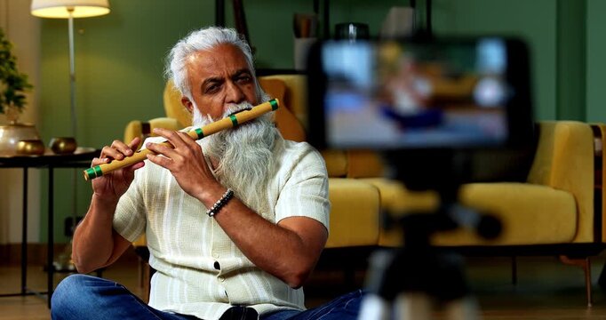 Indian mid age stylish bearded man teaching playing wooden flute musical instrument at home using smartphone, experienced classical bansuri tutor conducting online class in modern studio
