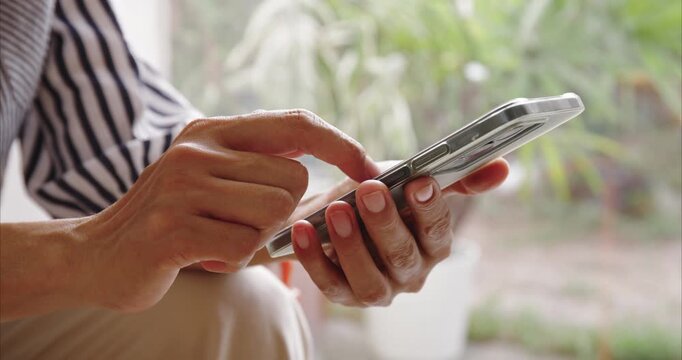 Close-up. Asian woman hands using smartphone on chair by window, scrolling screen with finger in bright home interior, mobile technology and digital lifestyle concept