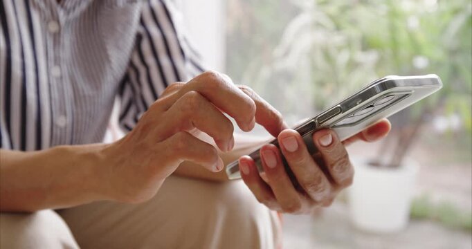 Close-up. Asian woman hands using smartphone on chair by window, scrolling screen with finger in bright home interio