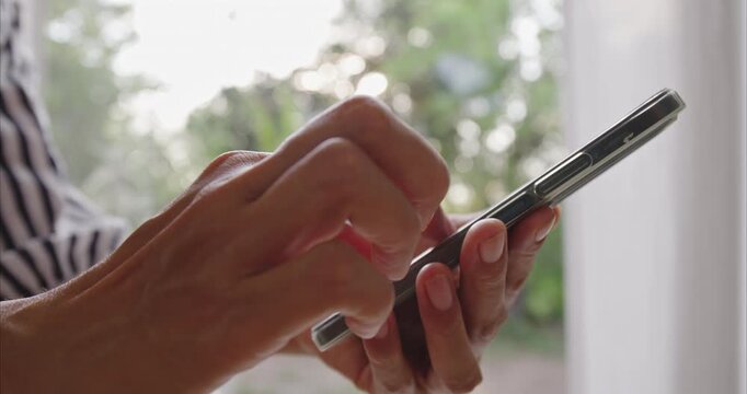 Close-up. Asian woman hands using smartphone on chair by window, scrolling screen with finger in bright home interior, mobile technology and digital lifestyle concept