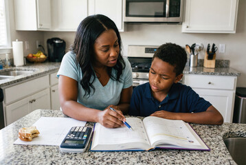 Una mujer ayuda a un estudiante con sus deberes de matemáticas en la cocina. Están concentrados en un libro de texto, con una calculadora y un refrigerio cerca en un ambiente hogareño y luminoso.