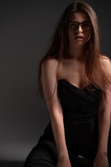 Naklejka premium Elegant young woman wearing eyeglasses and black outfit, sitting in a studio, posing with long brown hair, looking thoughtfully at camera with a mysterious gaze