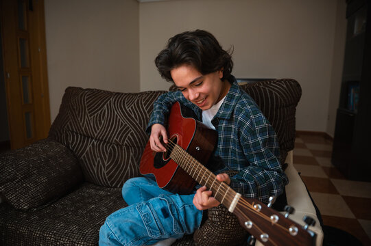 Teenager Boy Playing Acoustic Guitar At Home On Cozy Couch While Smiling And Strumming