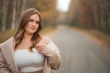 Obraz premium Young woman in a beige coat and white top posing on a road during autumn. plus size girl
