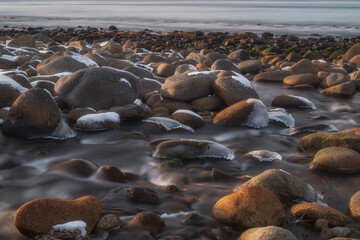 Long exposure photograph of snow-dusted boulders on a rocky shoreline with misty water flowing between them during winter at dawn.