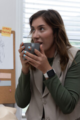 Adult woman holding gray mug, sipping and looking left in studio with corkboard, blinds, smartwatch