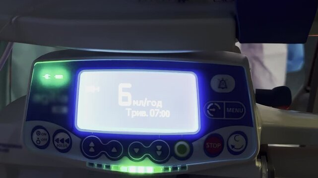 Medical infusion pump displays rate and volume on bright screen in ward. Device runs while indicator lights blink as therapy continues at procedure