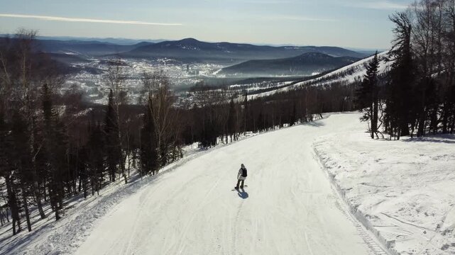 A drone view of a ski slope among tall trees, a ski piste at the Sheregesh resort. A snowboarder rides smoothly and softly on his board, evoking the atmosphere of freeriding