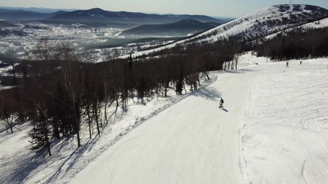 A drone view of a ski slope among tall trees, a ski piste at the Sheregesh resort. A snowboarder rides smoothly and softly on his board, evoking the atmosphere of freeriding