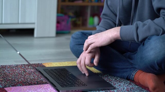 Male individual wearing  hoodie sits cross-legged on colorful carpet, interacting with laptop, fingers poised above keyboard, focused on screen in a cozy indoor environment