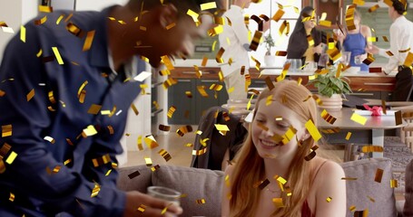 Smiling red-haired woman sitting on gray sofa in living room, taking plastic cup amid gold confetti