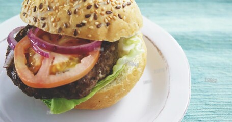 Showing sesame-seed hamburger sitting left on round white plate over teal-blue textured cloth © vectorfusionart