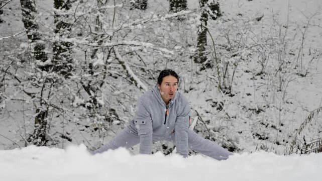 Man Training Split Snowy Clearing, Full Side Split On Snow With Hands Planted, Gray Hoodie, Face Showing Effort, Crisp Winter Trees Backdrop, Flexibility Challenge And Outdoor Exercise