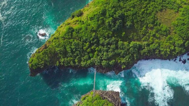 Top-down drone view of lush green coastal cliff surrounded by clear turquoise ocean water and crashing waves in tropical setting. Located in Kalong Island, Yogyakarta