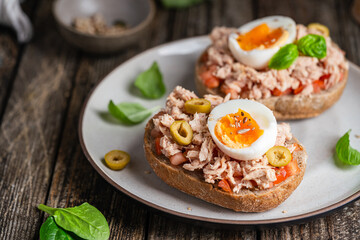 Tuna toast. Open sandwiches with whole grain bread, canned tuna, boiled egg, tomatoes and olives on wooden background. Healthy breakfast food