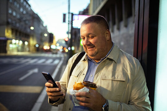 Smiling man with coffee using smartphone at city bus stop at dusk