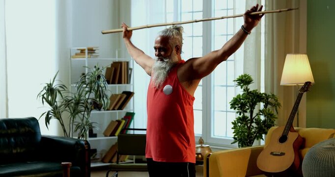 Indian mid age stylish bearded man doing exercise using stick, rod or staff at home, fitness conscious mature male performing strength and mobility workout showing agility and determination