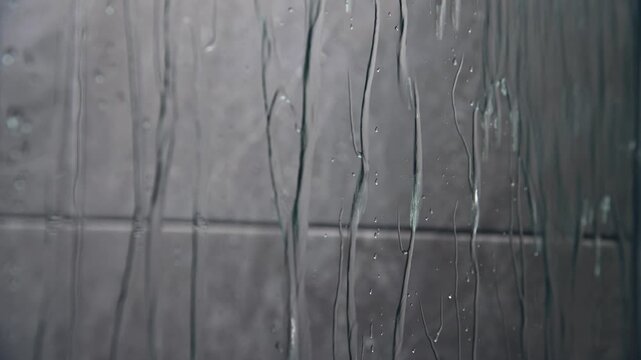 Water droplets cascade down a glass surface, creating a mesmerizing flow pattern against a textured gray wall in a bathroom setting