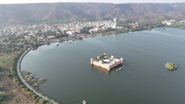 Aerial drone pull away from Jal Mahal historic water palace located in Man Sagar Lake in Jaipur, Rajasthan, India with city and mountains in background.