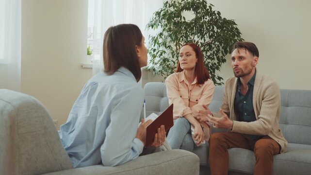 Emotional woman wiping tears during therapy session, sitting with partner while counselor listens and writes notes in notebook