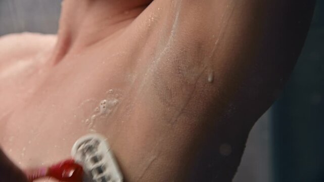 Female individual shaving in shower applying shaving cream to underarm with visible water droplets and soap bubbles, showcasing personal grooming routine in a bathroom setting