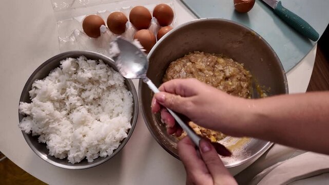 A woman cooks meatballs. A woman adds boiled rice to a metal bowl with minced meat
