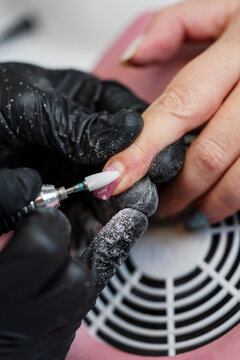 Close-up of a professional manicurist in black gloves using an electric nail file to remove old gel polish from a client's fingernails in a beauty salon setting.