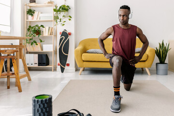 Naklejka premium Young adult black man performing lunges in living room, using exercise equipment for fitness and healthy lifestyle. Fitness concept