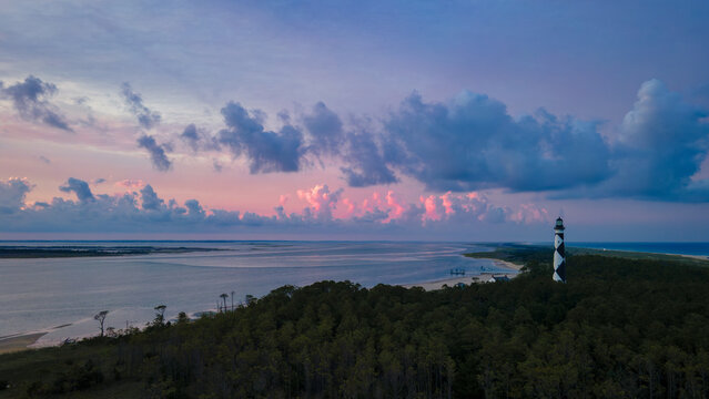 A soft dusk sky settles over Cape Lookout with pastel pink clouds glowing along the horizon. The calm channel below reflects hints of lavender and blue as it winds between the barrier islands. The