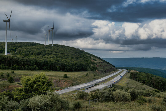 A line of wind turbines rises from the forested ridge in the Allegheny Mountains, standing against a dramatic sky of dark storm clouds and scattered light. Below, a winding mountain highway cuts