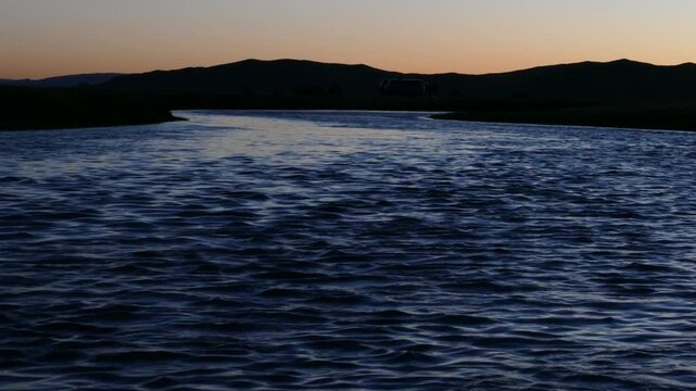A night river against the background of low mountains and a tourist camp under a pink sunset sky. Summer warm night.
