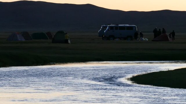 A evening river against the background of low mountains and a tourist camp under a pink sunset sky. Summer warm night.
