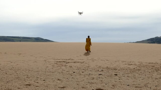 A Mongolian woman in a yellow national costume goes into the distance in the desert, above her flying quadcopter. Summer sunny day.