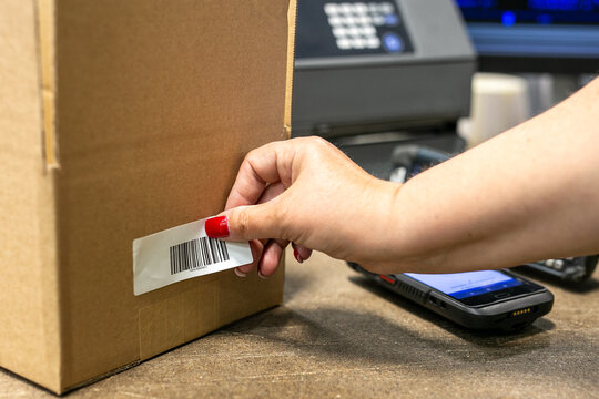 Warehouse operative applying a barcode label onto a cardboard box, with a label printer visible in the background.
