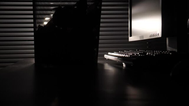 Desktop computer setup featuring a glowing monitor and keyboard on a desk, creating a silhouette effect against window blinds and reflecting the light from an open trash can