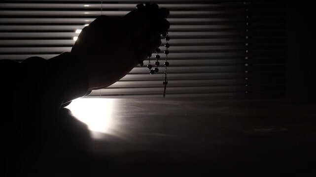 Gloomy hands holding a rosary with a cross against blinds, with light shining through, creating a strong silhouette symbolizing faith, solace, spirituality, and hope in difficult times