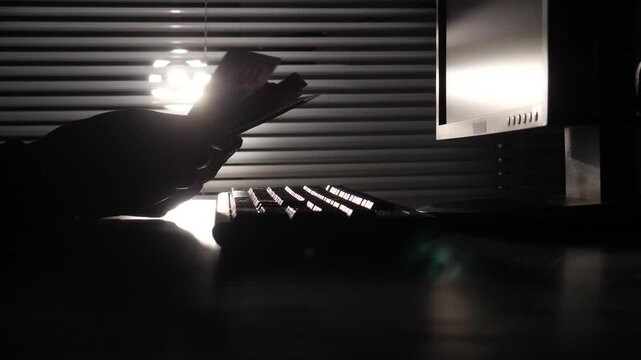 Close up silhouette of a cybercriminal counting cash while sitting at a computer desk, representing illegal financial activities and digital security threats