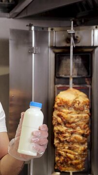 Chef, wearing an apron and gloves, showing a bottle of ayran as the best drink for kebab while standing by a vertical rotisserie in a restaurant kitchen