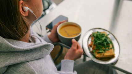 Young woman wearing headphones enjoys a meal of soup and sandwich while listening to music
