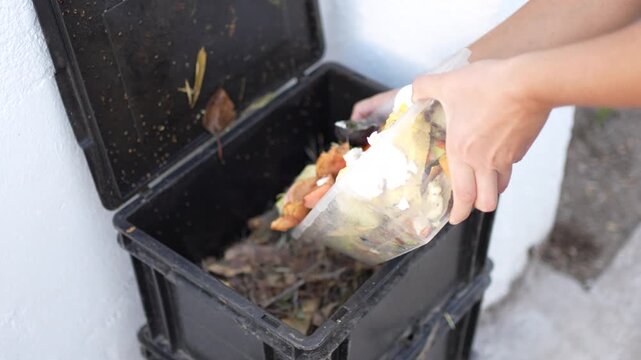 Close-up of a woman's hands throwing organic waste into a black recycled plastic urban compost bin.