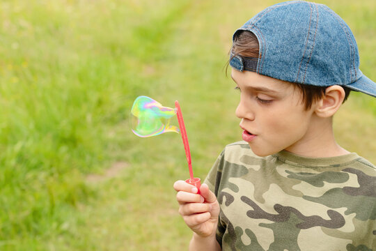 A boy is blowing a big soap bubble. He is wearing a blue cap. A khaki T-shirt. Green and yellow grassy background
