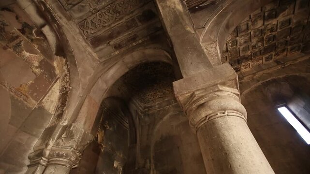 A low angle view panning across the dark, ancient stone interior of a historic monastery, revealing massive carved columns, elegant arches, and intricate patterned details on the ceiling.

