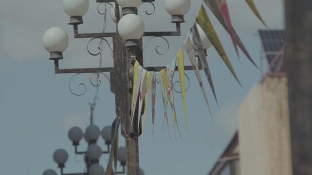 A string of decorative, colorful, triangular pennant flags with various unique illustrations gently flutters in the breeze against a blurred background of a city building and street lamps.

