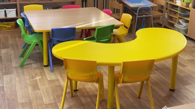 Interior of a kindergarten classroom. Small tables and chairs are arranged for children activities. Shelves with educational materials. The space is designed for early childhood learning. 